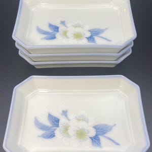 May include: Set of rectangular ceramic serving dishes. Each dish is white with a light blue trim and a floral design featuring white flowers and blue leaves. The dishes are stacked in two groups.