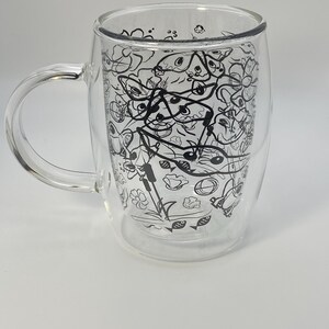 May include: A clear double-walled glass mug with a handle. The mug features a black and white doodle design of various shapes and objects. The mug is designed to keep beverages hot or cold.