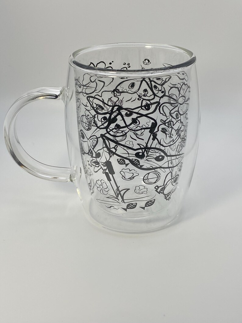 May include: A clear double-walled glass mug with a handle. The mug features a black and white doodle design of various shapes and objects. The mug is designed to keep beverages hot or cold.