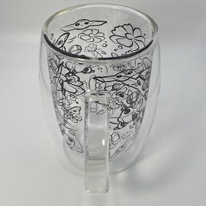 May include: A clear glass mug with a double-walled design. The interior features a black and white floral and abstract design. The mug has a clear handle and is suitable for hot or cold drinks.