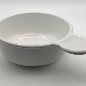 May include: A white ceramic soup bowl with a handle. The bowl is round with a slightly flared rim and a small, flat handle extending from the side. The handle is oval-shaped. The bowl is empty and sits on a white surface.