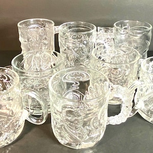 May include: A collection of clear glass mugs with embossed Batman-themed designs. The mugs feature various Batman characters and the iconic bat symbol. Each mug has a handle and a detailed, textured surface.
