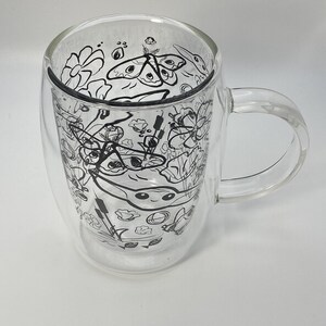 May include: Clear double-walled glass mug with a handle. The interior features a black and white line drawing of flowers, moths, and other botanical elements. The mug is designed to keep beverages hot or cold.
