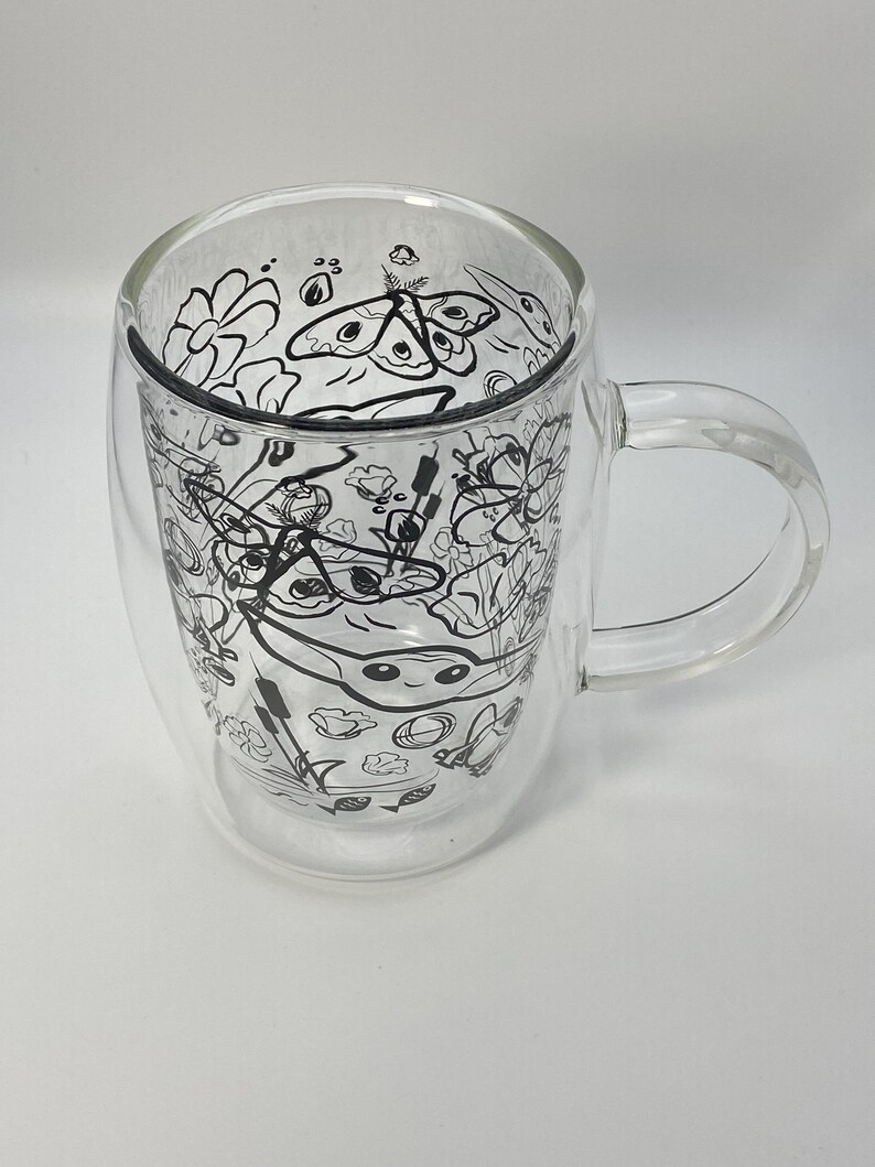 May include: Clear double-walled glass mug with a handle. The interior features a black and white line drawing of flowers, moths, and other botanical elements. The mug is designed to keep beverages hot or cold.