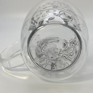 May include: Clear glass mug with a double-walled design. The mug features a handle and a decorative black illustration of fish, plants, and other aquatic elements. The mug is designed to keep beverages hot or cold.