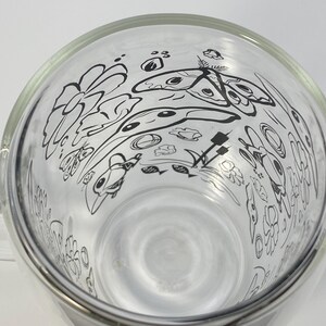 May include: A clear glass bowl with a wide rim, decorated with a black line art design of flowers, butterflies, and other organic shapes. The bowl has a smooth, reflective surface and is set against a white background. The design is visible from the inside.