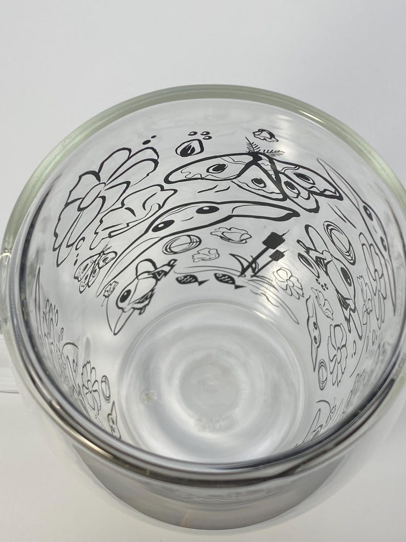 May include: A clear glass bowl with a wide rim, decorated with a black line art design of flowers, butterflies, and other organic shapes. The bowl has a smooth, reflective surface and is set against a white background. The design is visible from the inside.