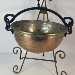 May include: A copper-colored metal cauldron with a black handle and a decorative stand. The cauldron has a rounded shape and is supported by a gold-colored metal stand with scrolled feet. The handle is attached to a decorative metal frame.