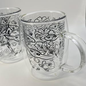 May include: Two clear glass mugs with a double-walled design. Each mug has a black and white doodle pattern of flowers, insects, and other whimsical elements. The mugs have a handle for easy gripping and are suitable for hot or cold drinks.