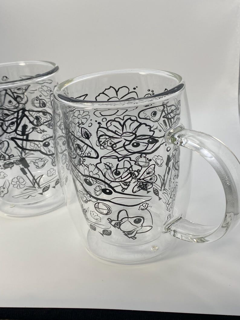 May include: Two clear glass mugs with a double-walled design. Each mug has a black and white doodle pattern of flowers, insects, and other whimsical elements. The mugs have a handle for easy gripping and are suitable for hot or cold drinks.