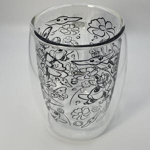 May include: A clear, double-walled glass with a black and white floral and creature design. The glass has a rounded shape and is designed to keep drinks hot or cold. The design includes flowers and cartoon-like creatures.