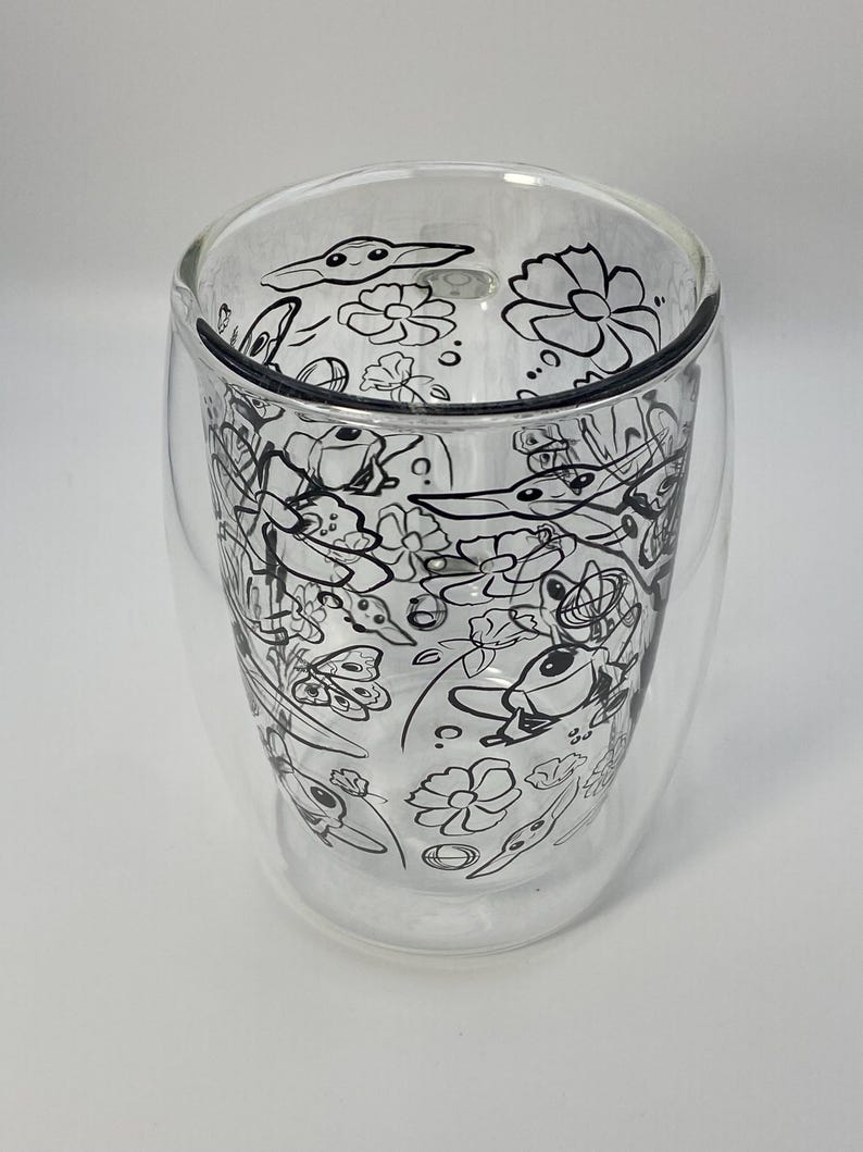 May include: A clear, double-walled glass with a black and white floral and creature design. The glass has a rounded shape and is designed to keep drinks hot or cold. The design includes flowers and cartoon-like creatures.