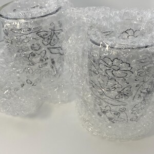 May include: Two clear glass tumblers with floral designs, wrapped in bubble wrap. The glasses have a delicate, etched pattern of flowers and leaves. The bubble wrap protects the glasses during transit.