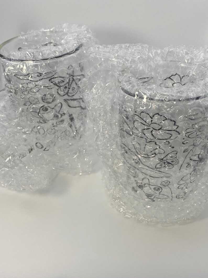 May include: Two clear glass tumblers with floral designs, wrapped in bubble wrap. The glasses have a delicate, etched pattern of flowers and leaves. The bubble wrap protects the glasses during transit.
