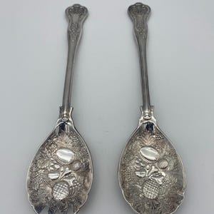 May include: A pair of silver-toned serving utensils consisting of a fork and a spoon. Each handle features an ornate, embossed design. The bowls of both utensils are decorated with detailed, raised relief patterns of various fruits and foliage.