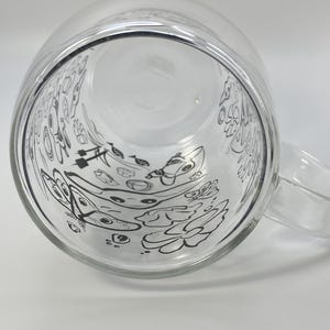 May include: A clear glass mug with a handle, decorated with black line drawings of flowers and other shapes on the inside. The mug is empty and clean.