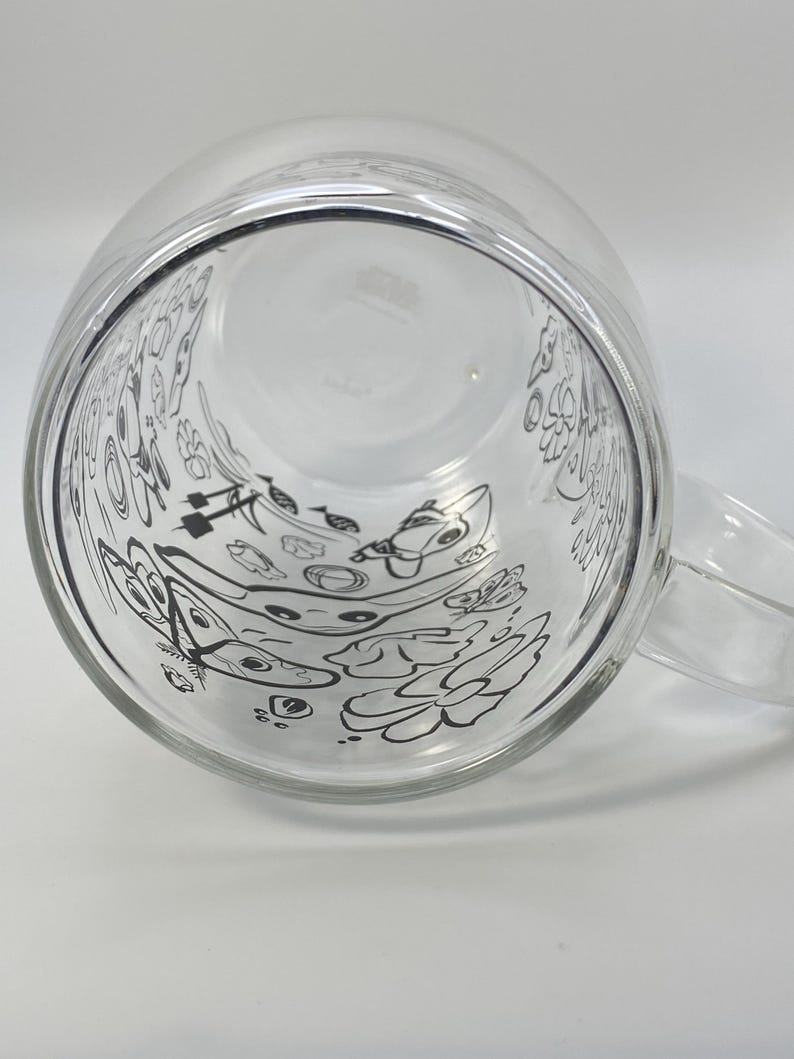 May include: A clear glass mug with a handle, decorated with black line drawings of flowers and other shapes on the inside. The mug is empty and clean.