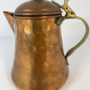 May include: A copper-colored metal teapot with a lid and a handle. The teapot has a spout for pouring and a decorative handle. The lid is attached with a hinge. The teapot is made of copper and has a shiny finish.