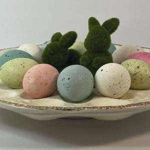May include: A decorative, off-white ceramic serving dish with a scalloped edge, filled with speckled pastel-colored Easter eggs and two green moss-covered bunny figures. The eggs are in shades of pink, blue, green, and white.