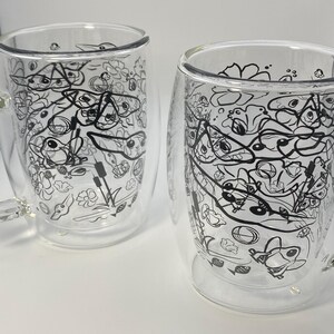 May include: Two clear glass mugs with a double-walled design. Each mug has a black and white doodle pattern of various shapes and creatures. The mugs are set against a white background.