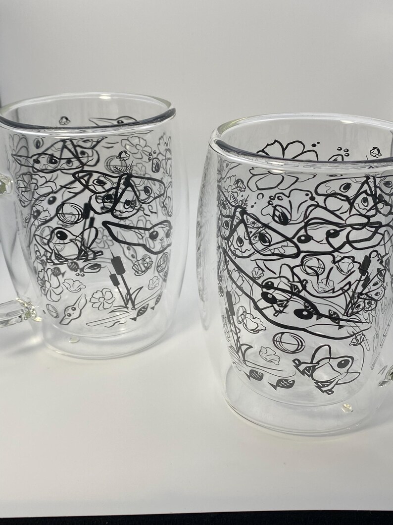 May include: Two clear glass mugs with a double-walled design. Each mug has a black and white doodle pattern of various shapes and creatures. The mugs are set against a white background.