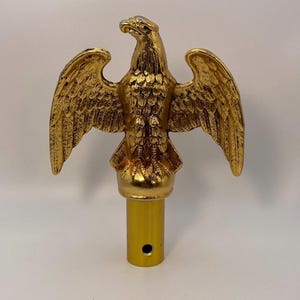 May include: A gold-colored eagle statue with outstretched wings. The eagle has detailed feathering and is mounted on a cylindrical base with a hole. The statue is likely a decorative element or a flag topper.