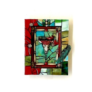 Puede incluir: Una obra de arte en mosaico que presenta una calavera blanca con flores rojas y una pluma turquesa. La calavera está enmarcada en un marco de mosaico rojo, blanco y verde.