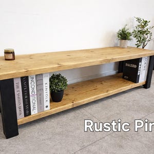 May include: A rustic pine bench with a natural wood top and a lower shelf. The legs are black. The bench holds books, a small potted plant, and a jar. The text "Rustic Pine" is at the bottom of the image.