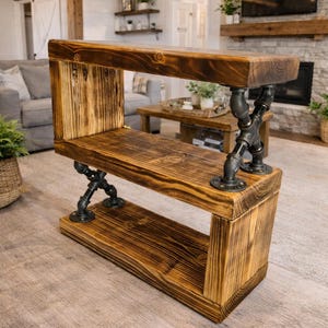 May include: A wooden console table with three shelves, supported by black metal pipe fittings. The wood has a warm, brown tone. The table is designed for display or storage, and is suitable for a living room.