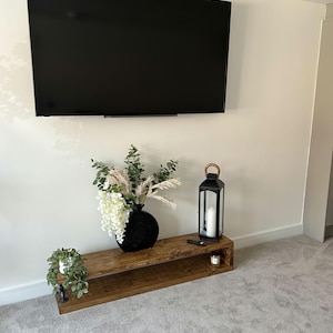 May include: A contemporary living room with a wall-mounted black television. Beneath it, a reclaimed wood media unit displays a black vase with flowers, a black lantern, and a small potted plant, creating a stylish, minimalist aesthetic.