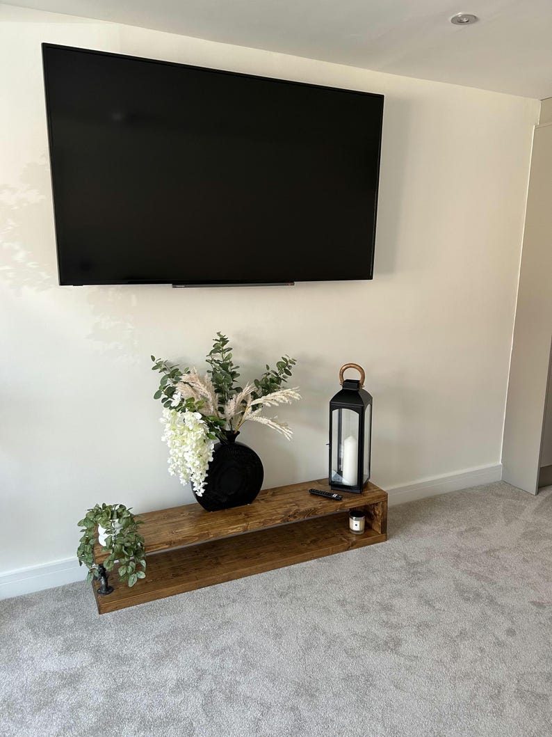 May include: A contemporary living room with a wall-mounted black television. Beneath it, a reclaimed wood media unit displays a black vase with flowers, a black lantern, and a small potted plant, creating a stylish, minimalist aesthetic.