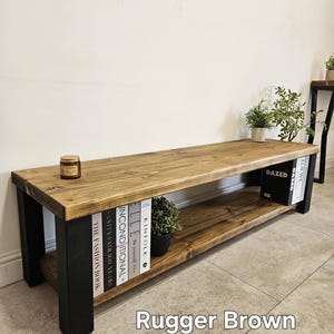 May include: A wooden media unit in Rugger Brown with black legs. The unit has a lower shelf and is styled with books and a small potted plant. A candle sits on the top surface. The unit is approximately 150cm long.