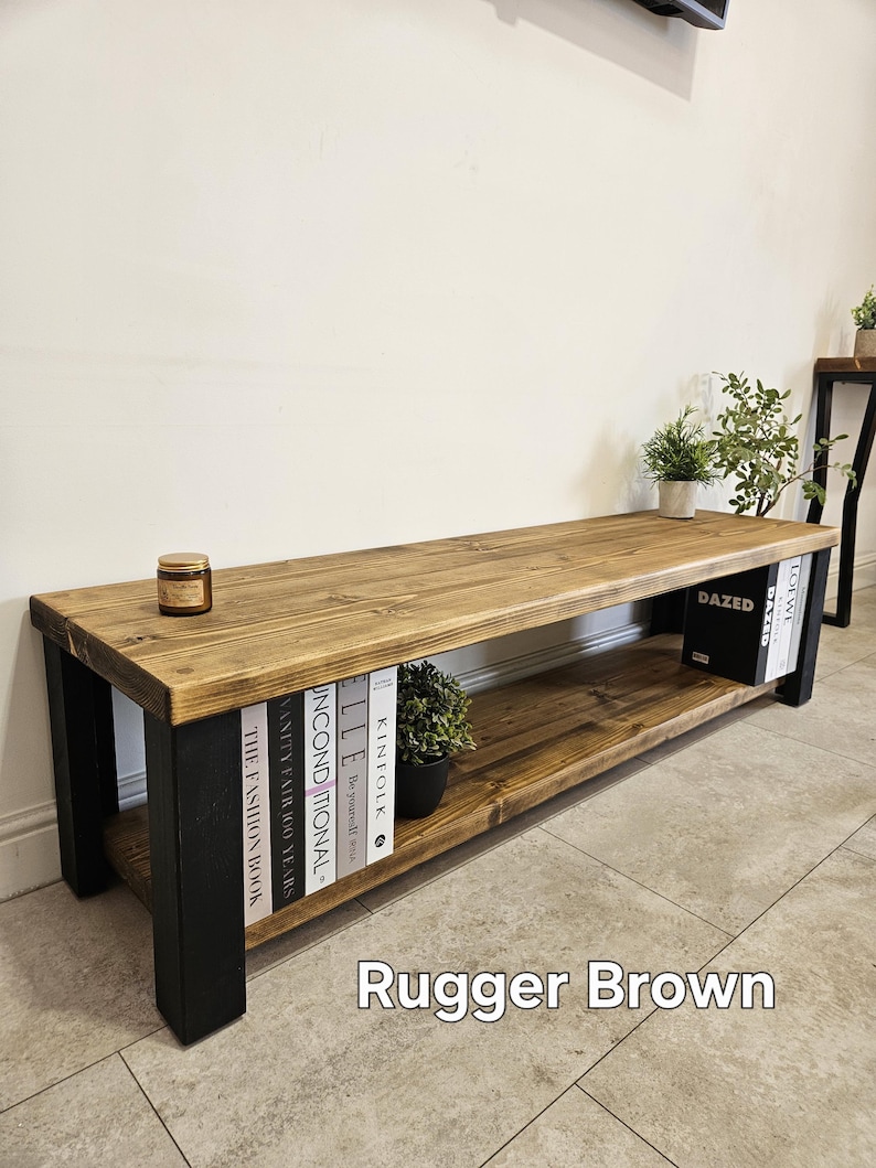May include: A wooden media unit in Rugger Brown with black legs. The unit has a lower shelf and is styled with books and a small potted plant. A candle sits on the top surface. The unit is approximately 150cm long.