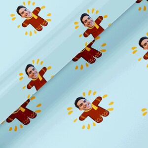 Puede incluir: Papel de regalo azul claro con un patrón repetido de hombres de jengibre con la cara de una persona. Los hombres de jengibre son marrones con detalles amarillos y una bufanda amarilla. El fondo tiene explosiones de estrellas amarillas.
