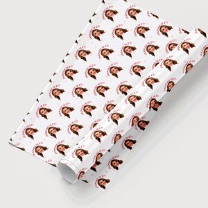Op de afbeelding: Wit cadeaupapier met een herhalend patroon van een vrouwen gezicht en de tekst "[Name] is 27!" in roze en rood.