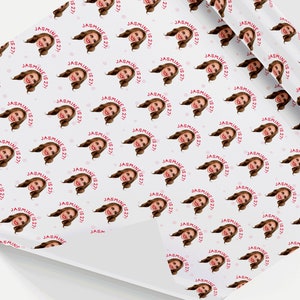 Op de afbeelding: Wit inpakpapier met een herhalend patroon van een vrouwengezicht in een cirkel met de tekst "JASMINE IS 27!" in roze. Er zijn kleine roze stippen verspreid over het papier.