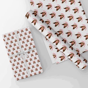 Op de afbeelding: Twee rollen wit inpakpapier met een herhalend patroon van een vrouwen gezicht en de tekst "Jasmine is 22!" in roze.