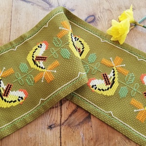 Op de afbeelding: Olijfgroene geborduurde tafelloper met een herhalend patroon van gele vogels, oranje accenten en groene bladeren. De randen zijn afgewerkt met een wit gestikte rand. Een paar gele narcissen rust op het houten oppervlak.