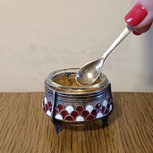 May include: A small, silver salt cellar with a glass top and a red and white enamel design. The cellar has three legs and is sitting on a wooden surface. A small silver spoon is resting in the cellar.