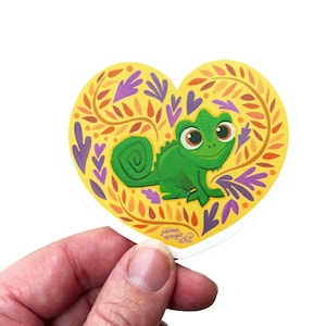 Puede incluir: Una pegatina en forma de corazón con un camaleón verde y un fondo amarillo con diseños de hojas naranjas y moradas. La pegatina se sostiene en una mano sobre un fondo blanco.