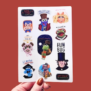 Op de afbeelding: Een stickervel met illustraties van Muppet-personages en -zinnen. De vellen bevatten afbeeldingen van personages als Miss Piggy, Kermit en Gonzo, samen met tekst als "Whatever" en "Hum Bug". De sticker wordt tegen een rode achtergrond gehouden.