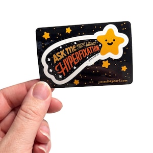 May include: A rectangular black sticker featuring a yellow smiling star and the orange text "ASK ME ABOUT MY LATEST HYPERFIXATION". The design includes small yellow stars and white sparkles against a dark background with the website jilliankayeart.com.