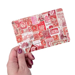 May include: A rectangular card with a collage of vintage postage stamps in red and pink hues. The stamps showcase diverse designs, including portraits and buildings. The card is held by a hand with painted nails.
