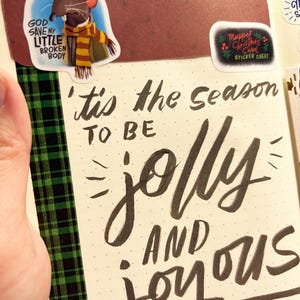 Op de afbeelding: Een journaalpagina met de zin "'tis the season to be jolly and joyous" in zwarte kalligrafie. Stickers zijn onder meer een Muppet-personage en een "Muppet Christmas Carol" sticker sheet. Groene en zwarte geruite tape accentueert de pagina.