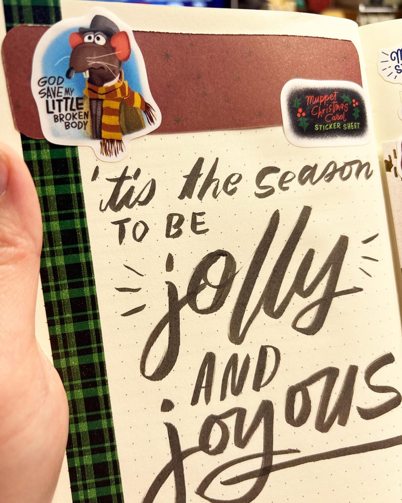 Op de afbeelding: Een journaalpagina met de zin "'tis the season to be jolly and joyous" in zwarte kalligrafie. Stickers zijn onder meer een Muppet-personage en een "Muppet Christmas Carol" sticker sheet. Groene en zwarte geruite tape accentueert de pagina.