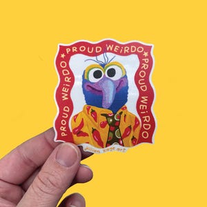 May include: A die-cut sticker featuring a purple, long-nosed character wearing a yellow shirt with a red chilli pepper pattern and a green tie. The character is framed by a red border with the text PROUD WEIRDO repeated and Jillian Kaye art at the base.