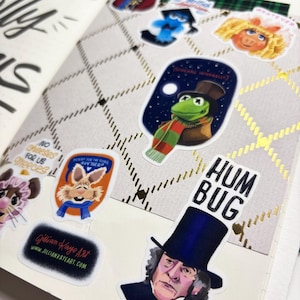 Op de afbeelding: Open notitieboekpagina met stickers van stripfiguren, waaronder Kermit de Kikker en Miss Piggy. De pagina heeft een ruitpatroon met gouden accenten en handgeschreven tekst. Een sticker luidt "HUM BUG".