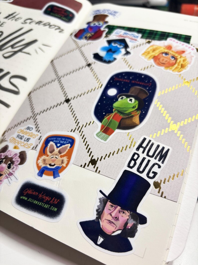 Op de afbeelding: Open notitieboekpagina met stickers van stripfiguren, waaronder Kermit de Kikker en Miss Piggy. De pagina heeft een ruitpatroon met gouden accenten en handgeschreven tekst. Een sticker luidt "HUM BUG".