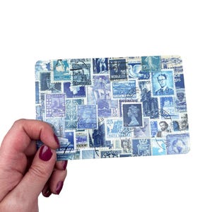 May include: A rectangular card featuring a collage of blue and white vintage postage stamps. The stamps depict portraits, buildings, and ships. The card is held in a hand with dark red nail polish. The stamps are from various countries.