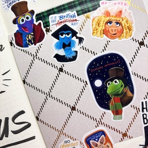 Op de afbeelding: Een verzameling kleurrijke stickers met Muppet-personages. De stickers bevatten Gonzo, Miss Piggy en Kermit, met teksten als "Mr. God-like Smarty-Pants" en "It is the British American Way!". De stickers bevinden zich op een patroonachtergrond.